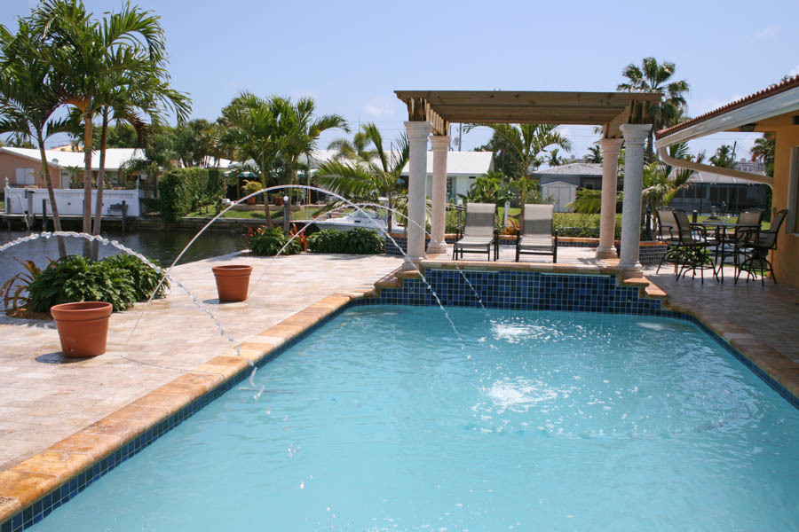 Pool with arcing deck jets, a columned pergola, and a travertine deck beside a canal